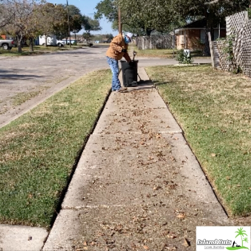 Worker from Island Cuts Lawn Service cleaning leaves from a sidewalk using a rake and garbage bin.